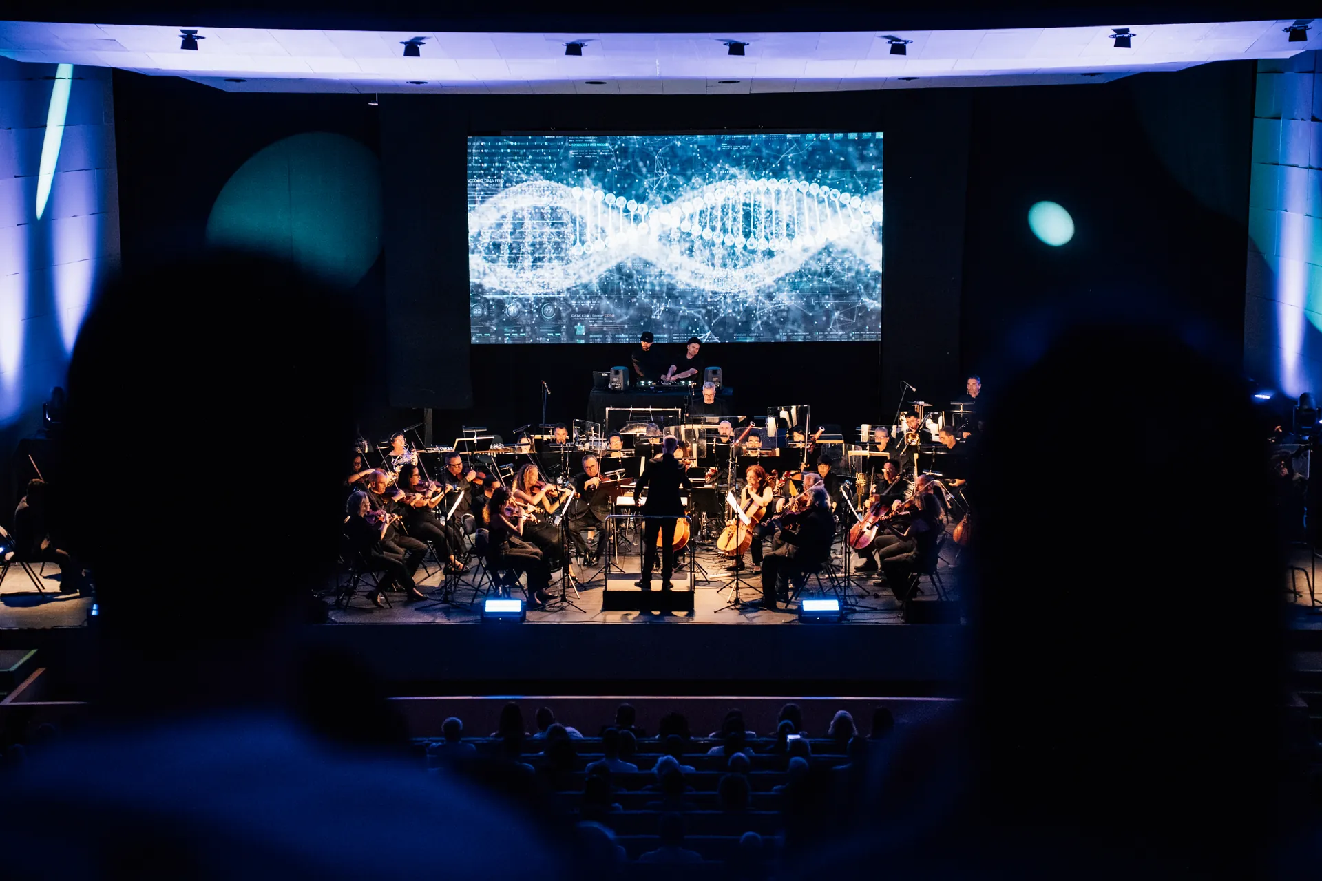 La orquesta y los DJs en escena bajo una gran pantalla, vistos desde el público